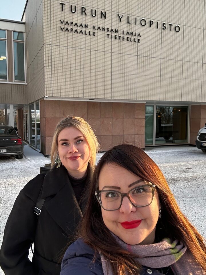 Tutkijat Jenny Paananen ja Kati Mäntysaari Turun yliopiston päärakennuksen edessä. Tutkijat hymyilevät. On talvi, ja taustalla näkyy lunta. Rakennuksen seinässä on teksti "Turun yliopisto. Vapaan kansan lahja vapaalle tieteelle".