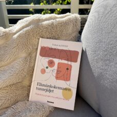 Kuvassa teos Elämänkokemuksen tunnejäljet on tuolin päällä aurinkoisella terassilla. Tuolilla on myös pörröinen viltti. Taustalla näkyy valkoinen rimakaide ja kaiteen takana vihreää pihaa.