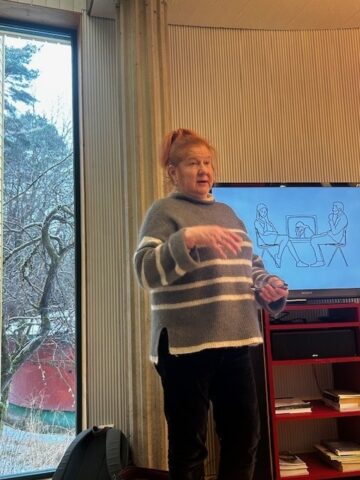 Psykoterapeutti Tarja Koffert luennoi suuren näytön edessä. Koffert elehtii käsillään puhuessaan. Taustalla on ikkuna, josta näkyy talvinen maisema.