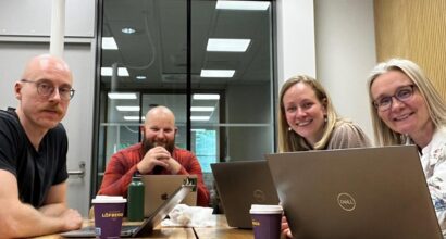 Picture of team members sitting around a table in a meeting room with laptops and coffee cups.