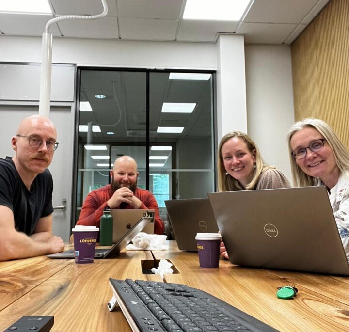 Picture of team members sitting around a table in a meeting room with laptops and coffee cups.