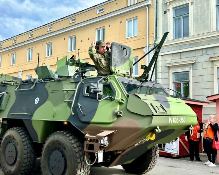 Maastokuvioinen panssarivaunu kadulla kerrostalojen edessä miehen seisoessa auki olevassa luukussa.
