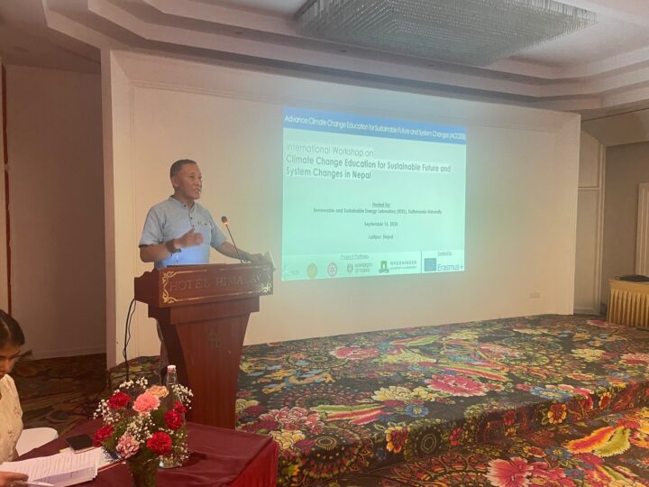 A man in a light blue shirt stands on a stage behind a podium, with the PowerPoint slide on the wall behind him saying "International Workshop on Climate Change Education for Sustainable Future and System Changes in Nepal". 