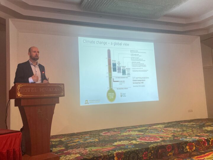 A man in a shirt and jacket stands on a stage behind a podium, the PowerPoint slide behind him on the wall says "Climate change - a global view" accompanied by some illustrations. 