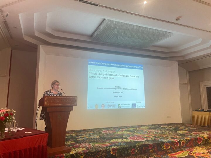 A spectacled woman stands on a stage behind a podium. The PowerPoint slide on the wall says "International Workshop on Climate Change for Sustainable Future and System Changes in Nepal".