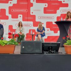 Paneelikeskustelu SuomiAreena-tapahtumassa: kolme henkilöä istuu lavalla pöytien ääressä ja yksi henkilö seisoo puhujapöydän takana, taustalla punavalkoinen SuomiAreena-logo ja lavan etureunassa punaisia pelargonioita kukkalaatikoissa.