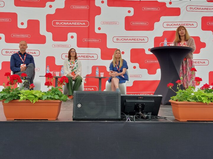 Paneelikeskustelu SuomiAreena-tapahtumassa: kolme henkilöä istuu lavalla pöytien ääressä ja yksi henkilö seisoo puhujapöydän takana, taustalla punavalkoinen SuomiAreena-logo ja lavan etureunassa punaisia pelargonioita kukkalaatikoissa.