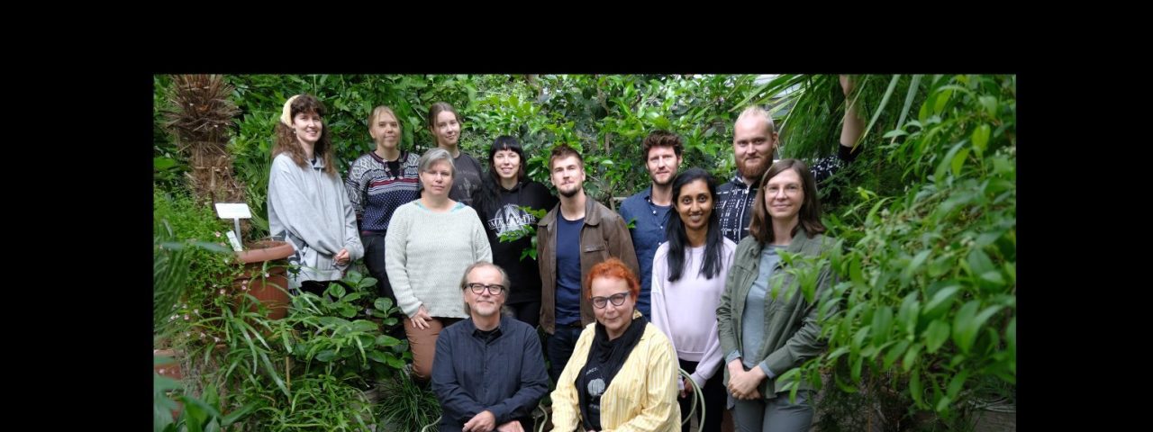 Group picture of the the HelanderSaikkonen Lab. Picture was taken in the Botanical gardens and Ruissalo, and the group of 8 women and 4 men are posing surrounded by foliage. The team members are standing around/behind the team leaders, who are seated.