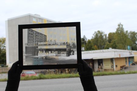 Rakennusta esittävä vanha valokuva, jota henkilö tarkastelee nykyhetken rakennusta vasten