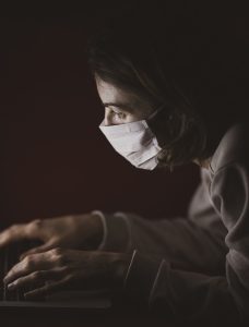 Nainen tuijottaa pimeässä läppärin ruutua maski kasvoillaan