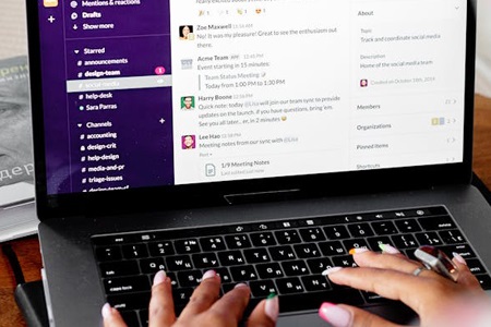 Henkilö kirjoittamassa kannettavalla tietokoneella, jonka näytöllä on viestintäsovellus avoinna eri työkanavineen ja keskusteluineen.