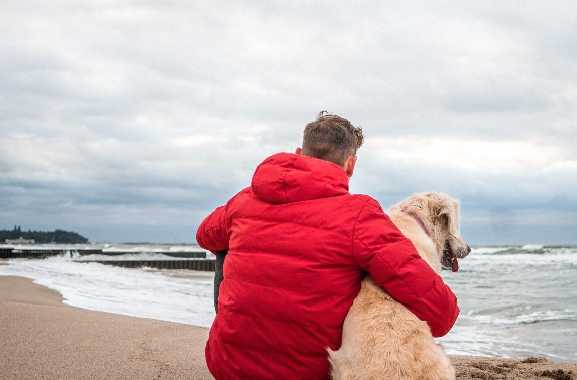 A red coated man sitting and hugging his white golden retriever on a beach watching the sea.