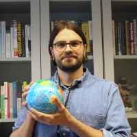 A brunette man in glasses holds a globe toy in front of a book shelf.