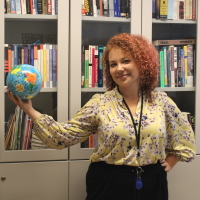 A ginger haired woman holds a globe toy in front of a book shelf and smiles.