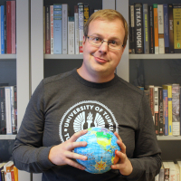 A man in a grey shirt holds a globe toy in front of a book shelf and looks straight ahead.