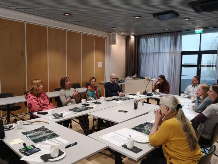 People sitting around a table. One person is speaking and the rest are listening attentatively and seem to be pondering the topic.