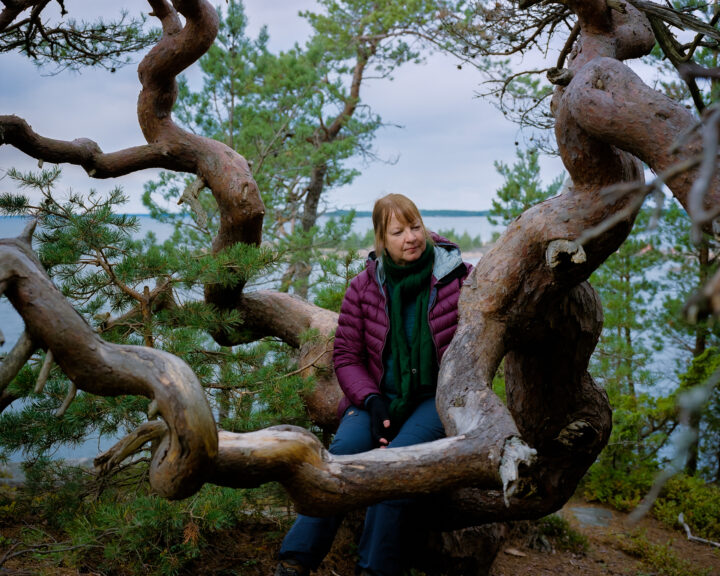 A woman is sitting on a fallen old tree trunk in a forest. Behind her is a few smaller trees and a view to a large open lake.
