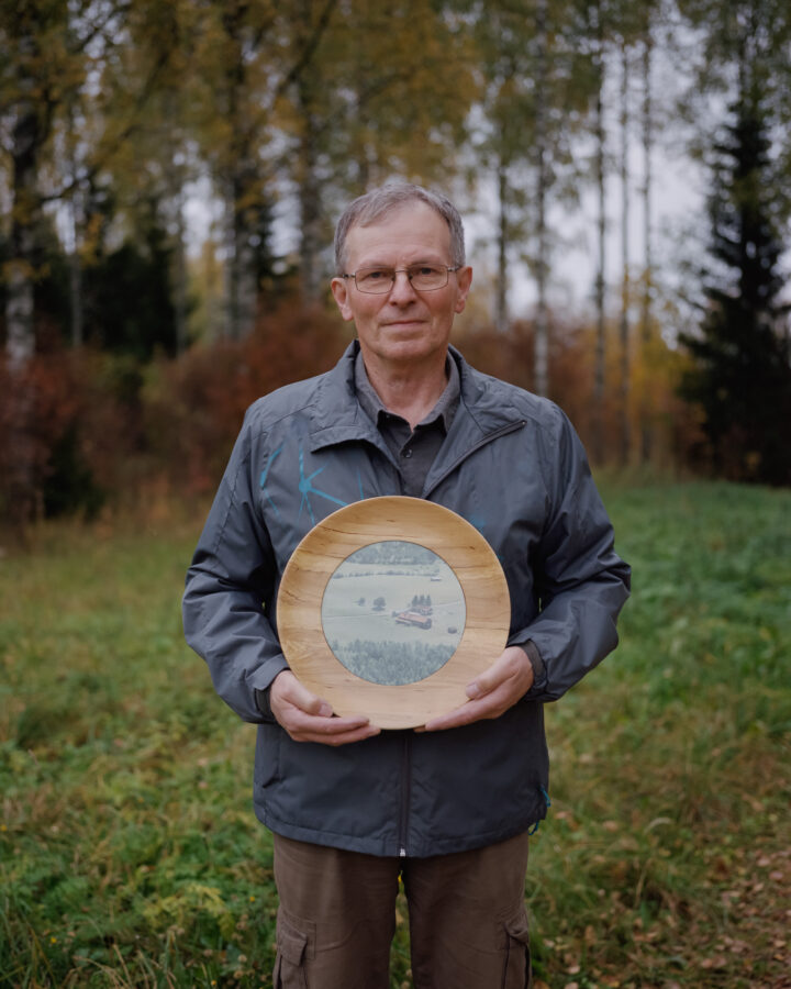 An elderly man is standing on a lawn with a round wooden plate on his hand. In the plate there is a photo of the same farm yard taken from an airplane many years ago. Behind him there is a forest.