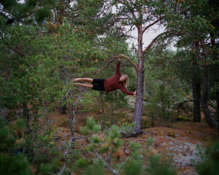 A man is in a forest. He is doing acrobatics using a small coniferous tree trunk to balance on.
