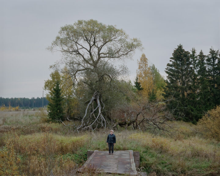An elderly woman is standing in front of a large decidous tree.
