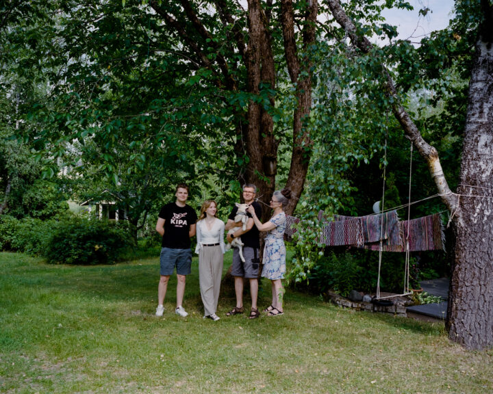 A family is standing in their yard in front of a big tree surrounded by lawn and several other trees.