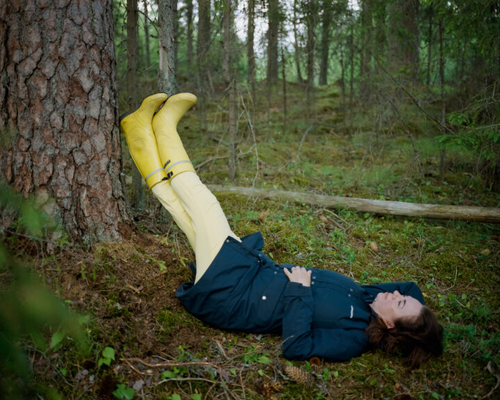 A woman is in a forest. She is lying on the moss resting her legs upwards leaning against a tree trunk.