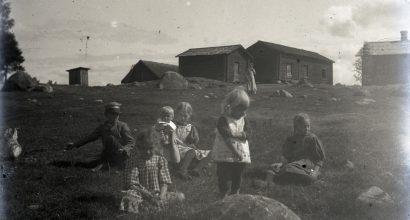 Kuusi alle 10-vuotiasta lasta leikkii heinikossa kesäisenä päivänä. Taustalla näkyy viisi hirsirakennusta ja pitkään mekkoon pukeutunut aikuinen.