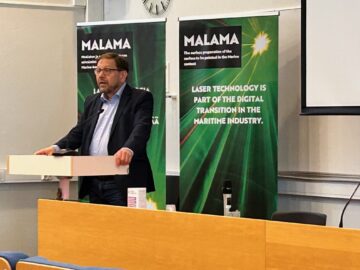 Tummaan pukuun pukeutunut mieshenkilö puhuu puhujapöntön takana. Taustalla kaksi vihreäsävytteistä rollup-telinettä.