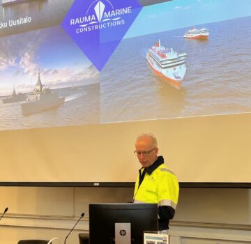 Keltaiseen takkiin pukeutunut mieshenkilö puhuu salin etuosassa taustalla Rauma Marine Constructionsin kalvo, jossa merellä laivoja.