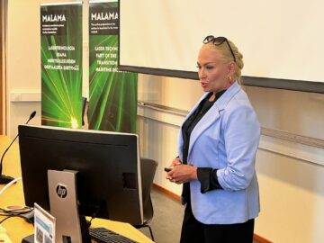 Siniseen bleiseriin pukeutunut vaaleahiuksinen naishenkilö puhuu valkokankaan edessä. Etualalla tietokoneen ruutu takaa ja taustalla kaksi pystytettyä vihreää rollup-telinettä.