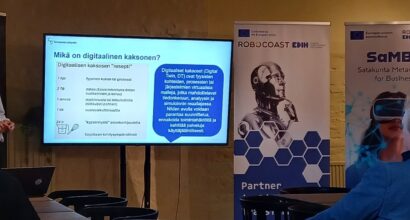 Esittelijä kertoo mitä digitaalinen kaksonen tarvitsee. Kaksi kuuntelijaa istuu kuuntelemassa. Taustalla Robocoastin ja SaMBa-hankkeen roll upit.