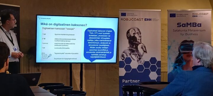 Esittelijä kertoo mitä digitaalinen kaksonen tarvitsee. Kaksi kuuntelijaa istuu kuuntelemassa. Taustalla Robocoastin ja SaMBa-hankkeen roll upit.