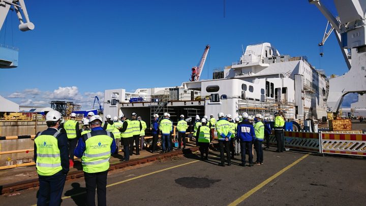 Lots of people with yellow vests in front of a ship in a shipyard.