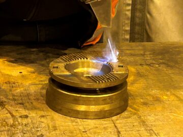 Pyöreää metallikappaletta työstetään lasersäteellä, josta nousee hieman savua.