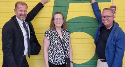 Kaksi miestä ja yksi nainen seisovat keltaisen seinän vieressä ison numero viitosen edessä. Molempien reunolla seisovien miesten toinen käsi on nostettu ylös High five -tyylisesti.