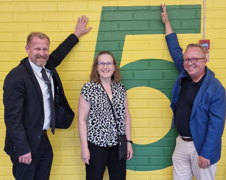 Kaksi miestä ja yksi nainen seisovat keltaisen seinän vieressä ison numero viitosen edessä. Molempien reunolla seisovien miesten toinen käsi on nostettu ylös High five -tyylisesti.