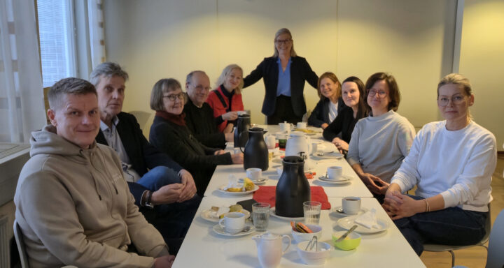 Hymyileviä ihmisiä kahvipöydän ympärillä katse kohti kuvaajaa.