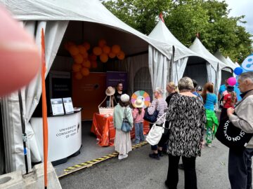 Vaalean harmaita telttoja vierekkäin katukuvassa, joissa etummaisessa joukko ihmisiä teltan edessä, esitteijä pöydän takana ja katossa oransseja ilmapalloja.
