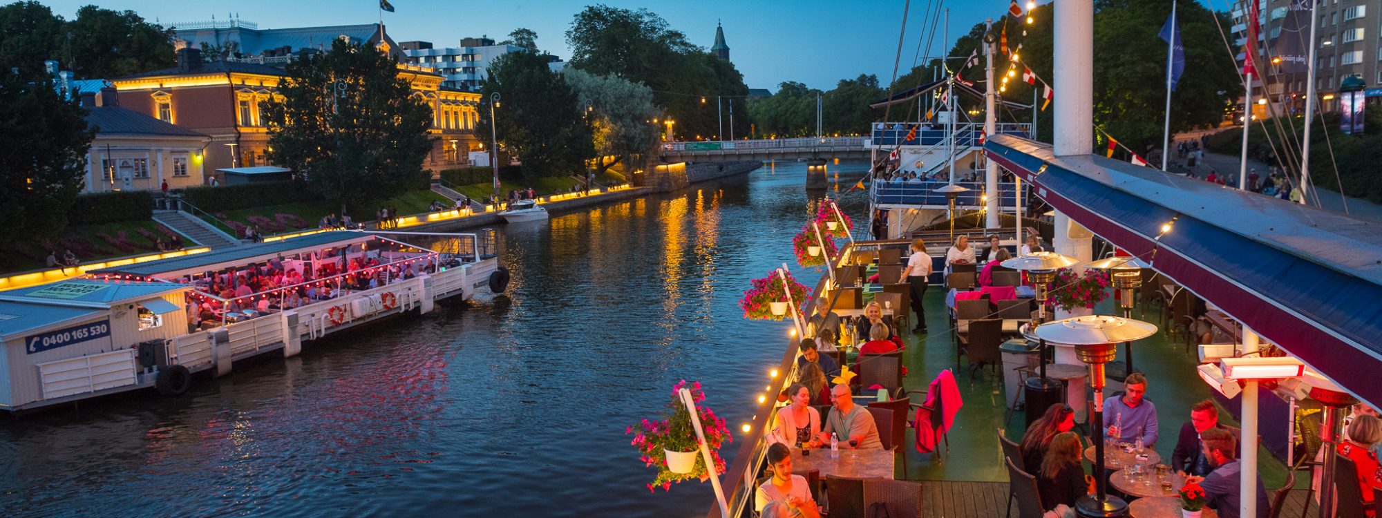 An evening photo of the Aura River in Turku. On both sides of the river there are lit up river boats with people enjoying beverages.