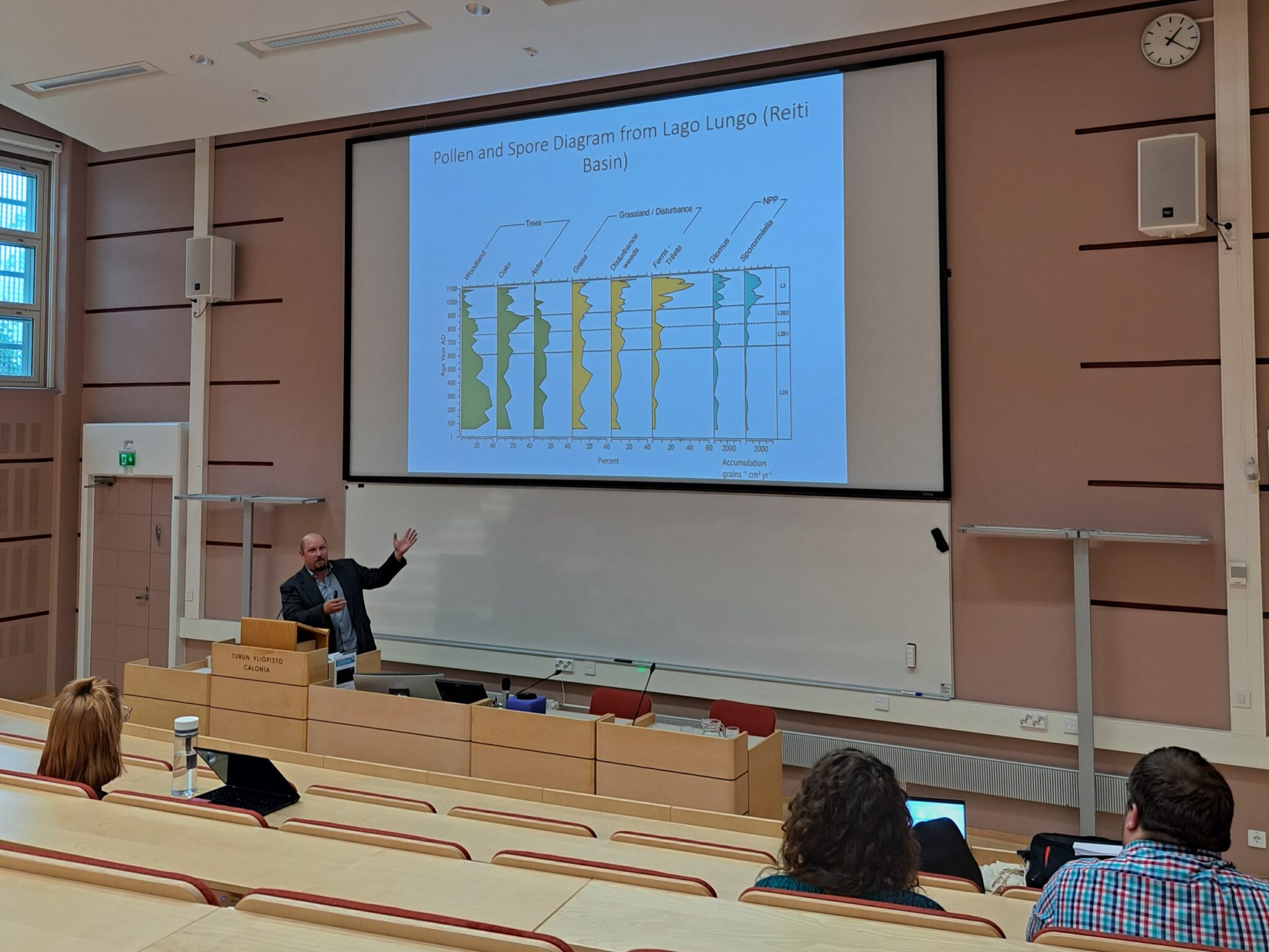 A person talking in the front of a lecture hall, with a slide show behind him.