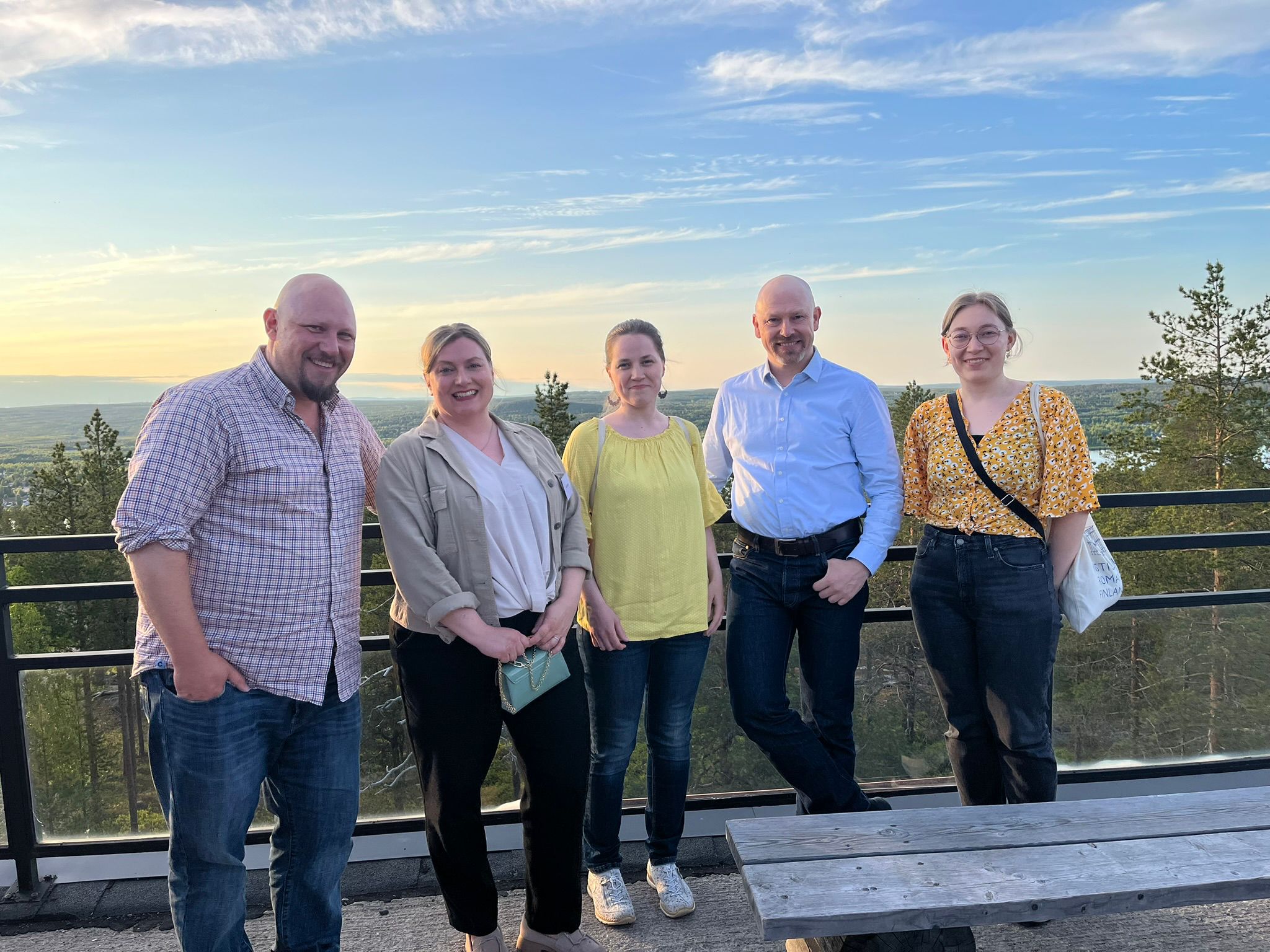 Five people standing in a row, with a forested landscape and blue sky behind them.