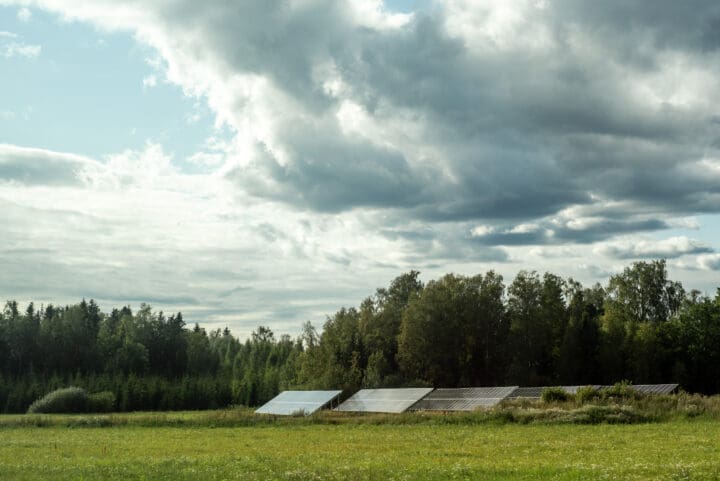 Aurinkopaneeleja riveissä pellolla, joka rajautuu metsään. Pellon päällä näkyy selkeä taivas ja jonkin verran raskaita pilviä.