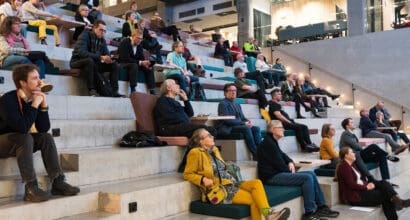 Yleisöä istumassa portailla katsomassa yläviistoon näytölle ja kuuntelemassa tutkijoiden esityksiä.