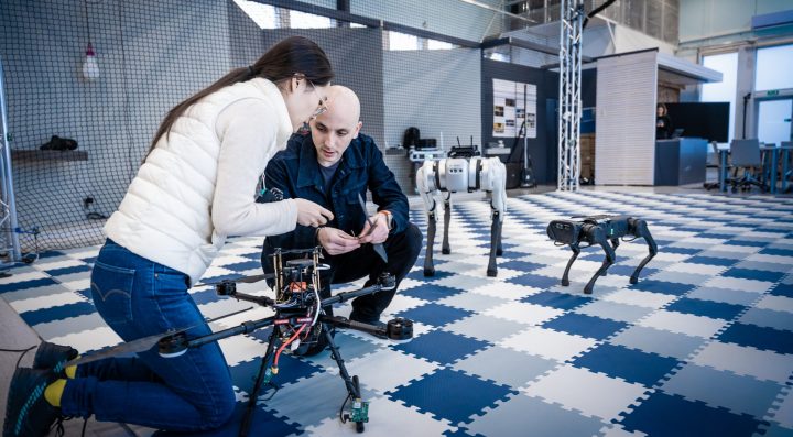 Two people fixing a drone with two robots in the background.