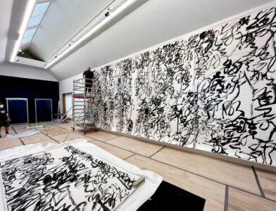 Gallerian työntekijä kiinnittää teoksen seinälle ylhäällä telineillä. Teoksen korkeus on 2 kertaa ja leveys 5 kertaa työntekijän pituus.